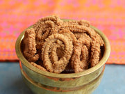 A brass bowl filled with Kambu Pepper Murukku, a traditional Indian snack, displayed against a colorful fabric background.